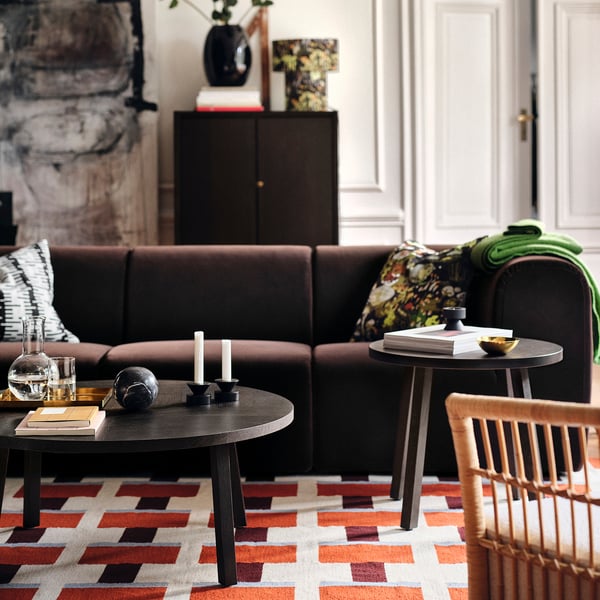 Elegantna dnevna soba z velikim rjavim kavčem, leseno omarico, mizicami in živahno preprogo STOCKHOLM 2025, ki pokriva večino tal.
