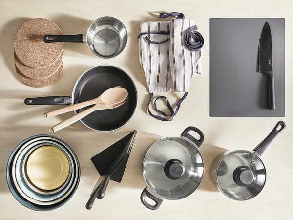 Kuchynské náradie zahŕňa hrnce, panvice, dosku na krájanie, nôž a utierky z nehrdzavejúcej ocele, úhľadne usporiadané na béžovom povrchu.