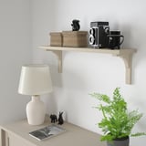Polica BERGSHULT v béžovej farbe pojme koše, fotoaparát, hrnček, dekor. Svetlé drevo, minimalistický dizajn nad stolom s lampou a rastlinami.