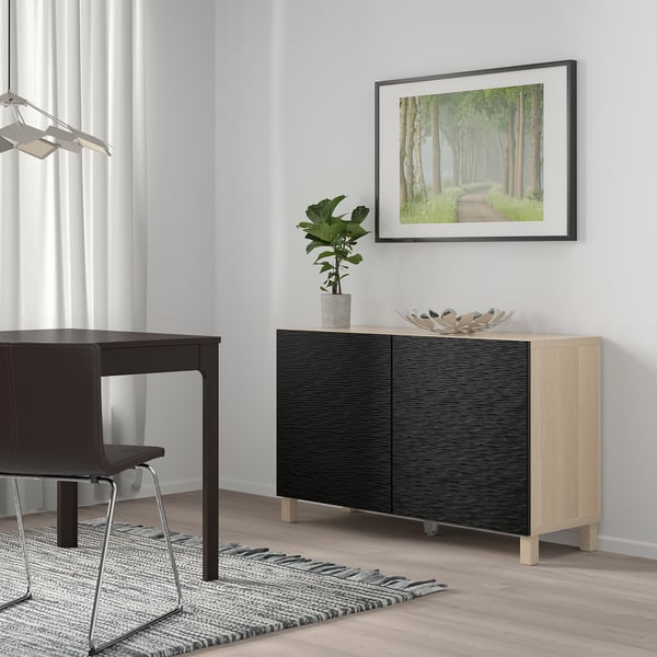 Minimalistická izba s nábytkom z čierneho a svetlého dreva. BESTÅ príborník, lesné umenie, tmavý stôl, rastlina, pruhovaný koberec.