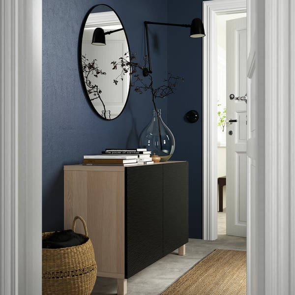 Moderná tmavomodrá predsieň s príborníkom BESTÅ, zrkadlom, tkaným košíkom a sklenenou vázou s konármi.