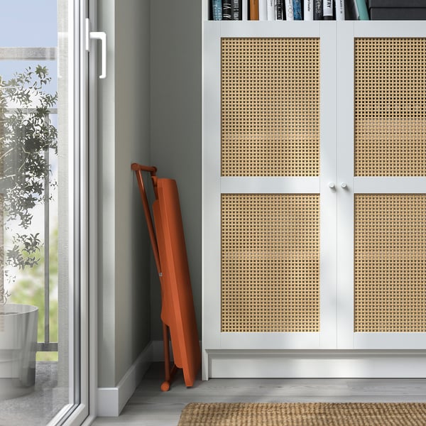 Skladací oranžový stojan na notebook sa opieral o biele skrinky s tkanými dverami, blízko okna s výhľadom na stromy.