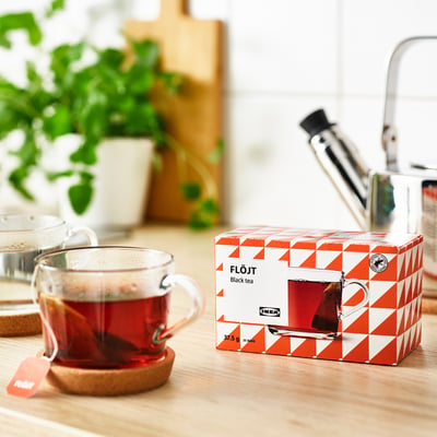 Priehľadný hrnček pariaceho sa čierneho čaju vedľa otvorenej krabičky na čaj IKEA FLÖJT s 25 čajovými vrecúškami na drevenom stole, čo naznačuje, že je pripravený na pitie.
