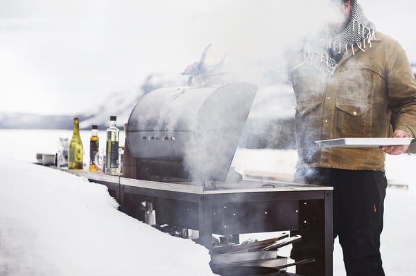 Muž grilujúci v zasnežený deň pomocou strieborného grilu GRILLSKÄR so stúpajúcou parou, držiaci biely tanier, obklopený fľašami.