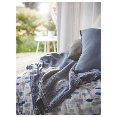 Modro-sivá deka na posteľ s geometrickým poťahom. Mäkký, recyklovaný polyester. Biela stolička a rastliny videné cez okno, ktoré vytvárajú útulnú atmosféru.