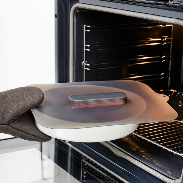 Osoba používajúca chňapky vkladá do rúry bielu misku s hnedým silikónovým vekom, aby zabránila rozstrekaniu a udržala jedlo čerstvé.