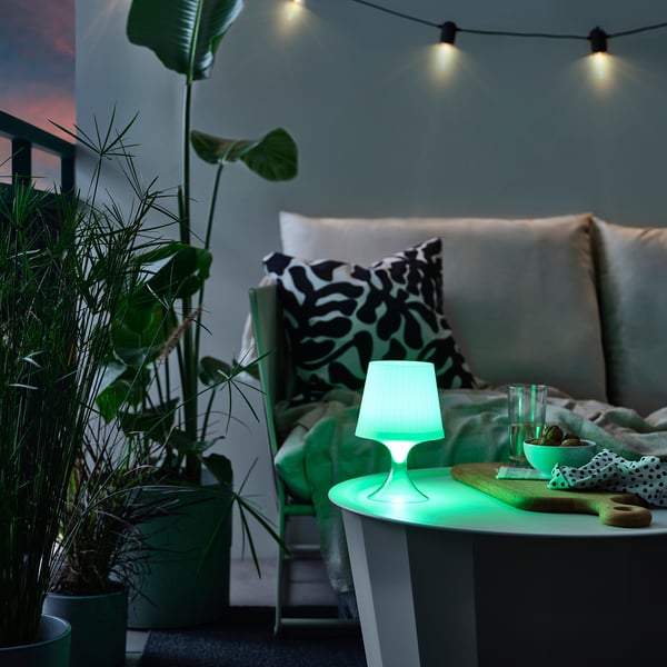 Malá zelená lampa LAMPAN na stole s rastlinami a gaučom za súmraku.