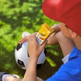 Dieťa pije žlté lang lur smoothie, s futbalovou loptou, v červenej čiapke.