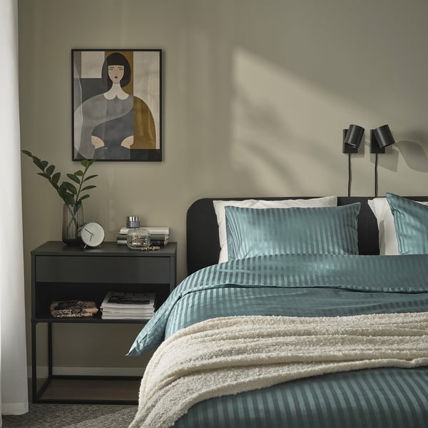 Moderná spálňa s modrozelenou posteľnou bielizňou NATTJASMIN, elegantným príručným stolíkom, nástennými umeleckými dielami a rastlinou. Mäkká, pruhovaná, saténová látka, úhľadne ustlaná posteľ.