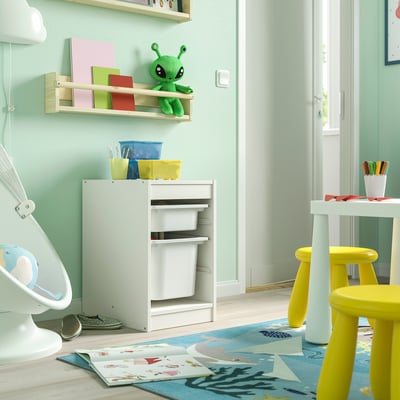 Detská izba s úložným priestorom TROFAST v bielej farbe, do ktorej sa zmestia farebné koše. Je nízky, obdĺžnikový, s tromi zásuvkami. V blízkosti je svetlý koberec a žlté stoličky.