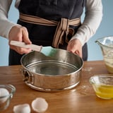 Človek používa zelenú kefu na vymastenie pružinovej formy z nehrdzavejúcej ocele obklopenej prísadami na pečenie.