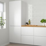Moderná kuchyňa s veľkou bielou chladničkou a skrinkami VOXTORP, elegantnými rukoväťami, svetlým pozadím, rastlinami. Vysoký, minimalistický.