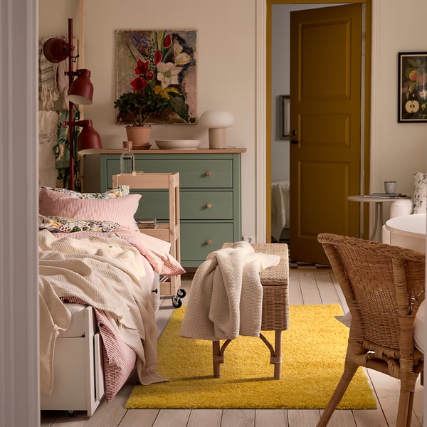 Bright yellow ÄRENDE rug in cosy bedroom, surrounded by white bed, wicker chair, and green dresser, adding warmth and sound absorption.