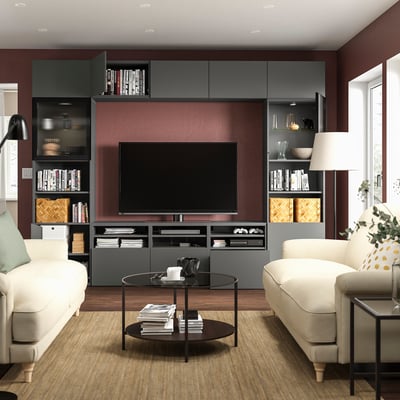 Modern living room with BESTÅ tv unit, white sofas, wooden tables, and a large flat-screen tv. Red wall, grey shelves, and stylish decor.