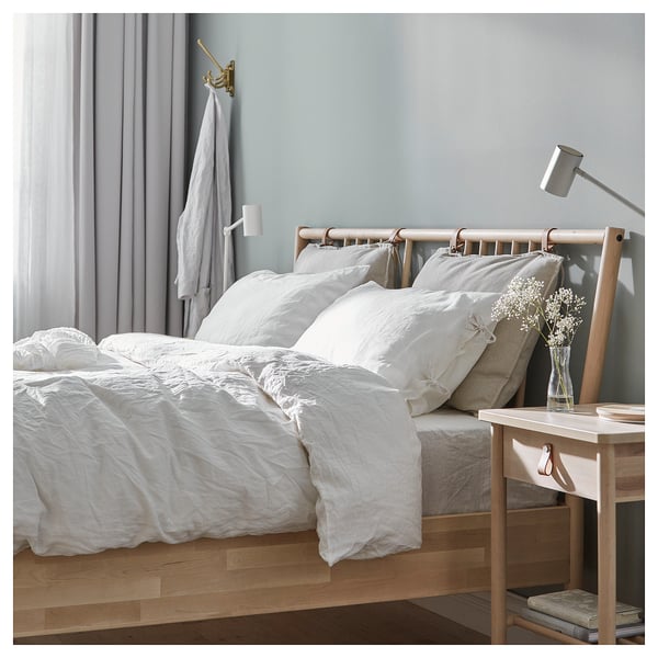 Minimalist bedroom scene with white bedding, light wooden bed, grey walls, and small side table with lamp and vase.