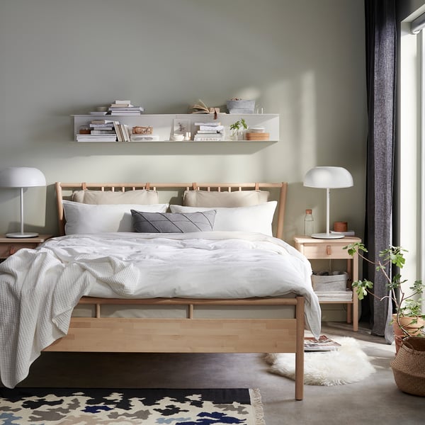 Modern bedroom with light wood BJÖRKSNÄS bed, white linens, grey pillows, nightstands, lamps, shelves, plants, and rug.