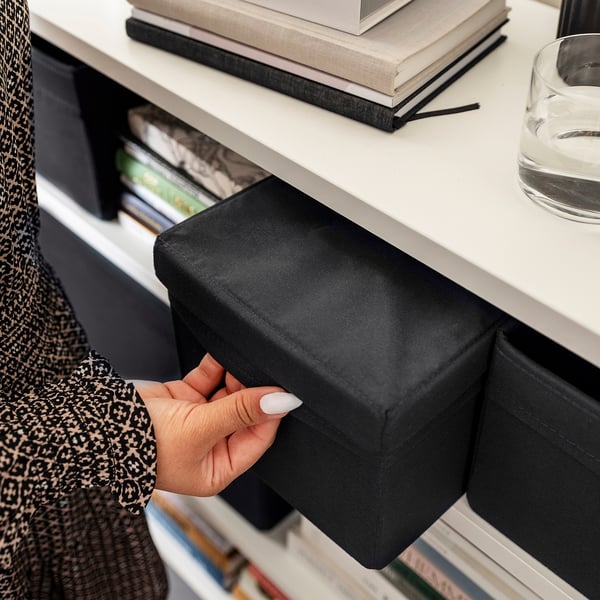 Hand pulls black fabric storage box from white shelf, containing various items, showcasing organisational functionality.