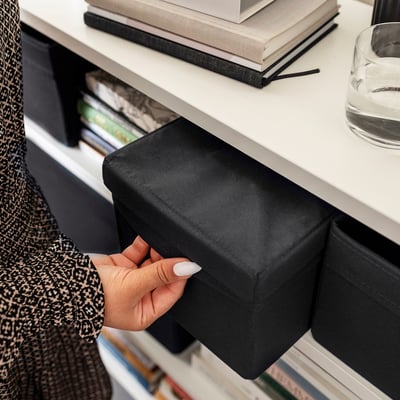 Hand pulls black fabric storage box from white shelf, containing various items, showcasing organisational functionality.