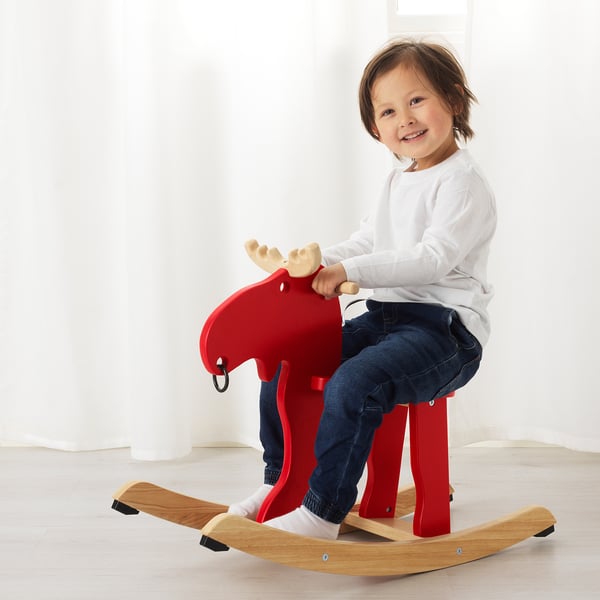 A young child sits on a red rocking moose toy, the mooses antlers are beige. The childs feet touch the wooden base. The child is smiling.