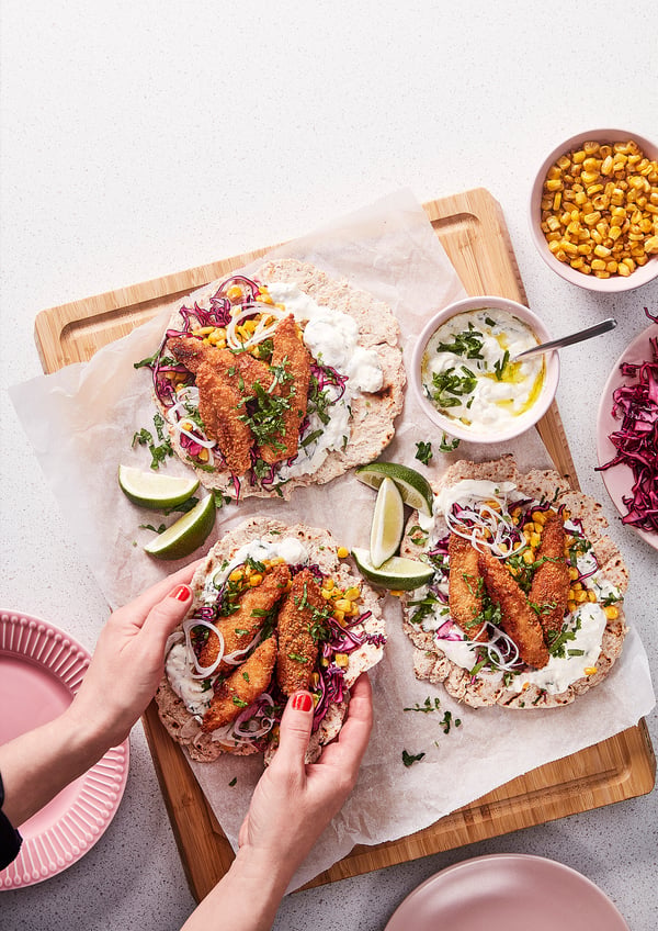 Two crispy fish tacos on a wooden board, topped with corn, lime, and herbs. A hand holds one taco, with additional toppings and plates nearby.