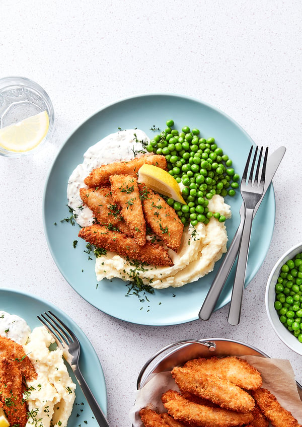 A plate of crispy golden fish bites on creamy mash with green peas and lemon. Two forks and a glass of lemonade on the side.