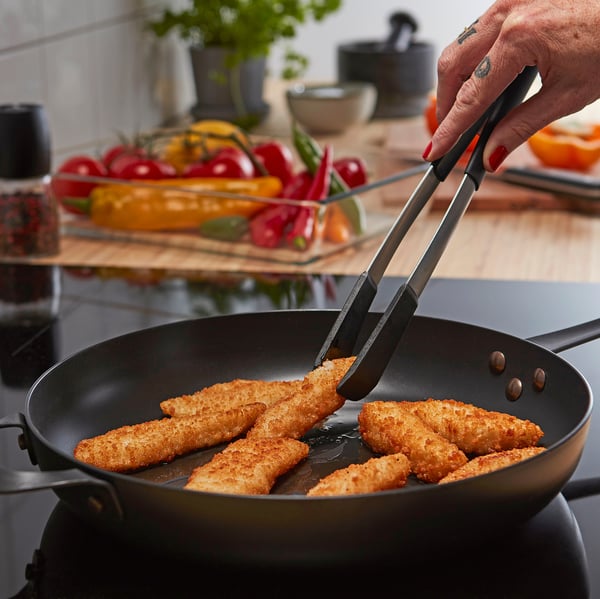 Person cooking breaded fish in a black frying pan, holding tongs, surrounded by colourful vegetables.