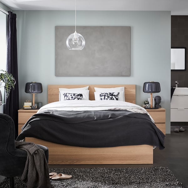 Stylish modern bedroom a light wooden honeycomb-patterned bed with grey linens, black lamps, large art, and industrial pendant lighting.