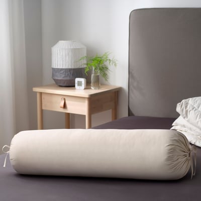 Bedroom with cylindrical bolster pillow on grey bed, beside table with lamp, clock, and plant.