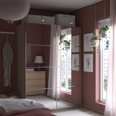 Modern bedroom with burgundy walls, featuring a reflective PAX wardrobe, hanging plants, and a window with sheer curtains.