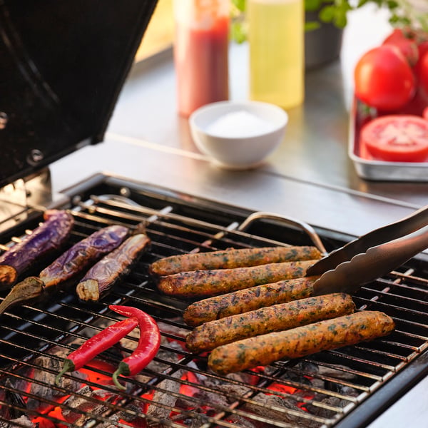 Image displays a grill with sausages and vegetables cooking. Sausages are brown, grill has tongs.
