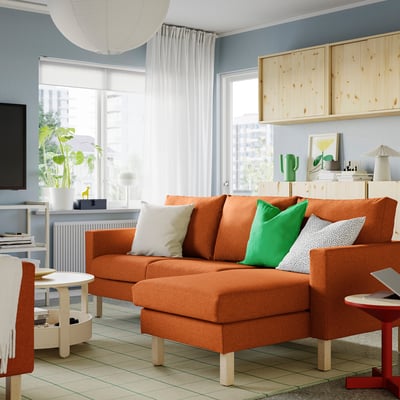 Bright living room with large orange SALTSJÖBADEN sofa, green pillows, wooden shelves, and a wide window.