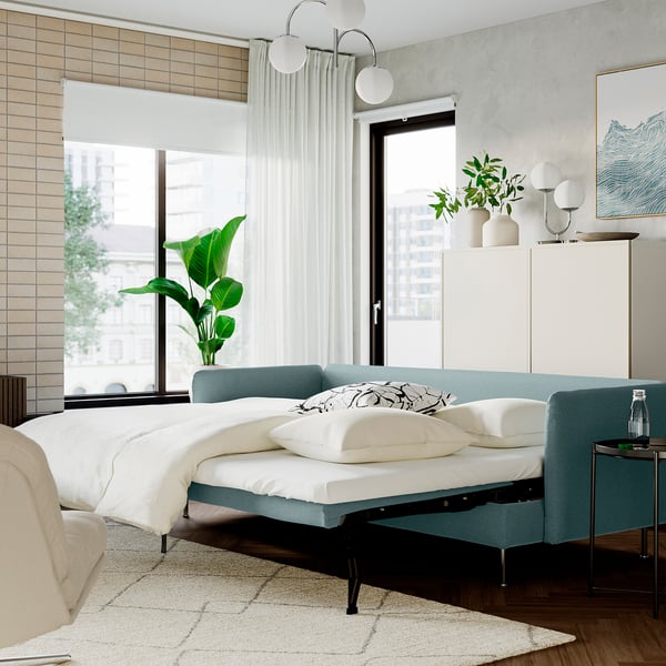 Modern bedroom with teal sofa-bed, white linens, big window, sheer curtains, cabinet, lights, and plant.