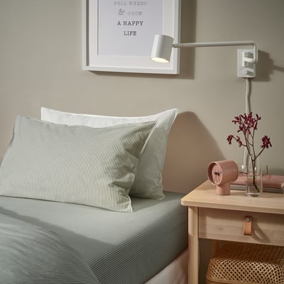 Bed with white and grey pillows, green striped sheets. Small wooden bedside table with small clock and plant. Picture with quote above bed.