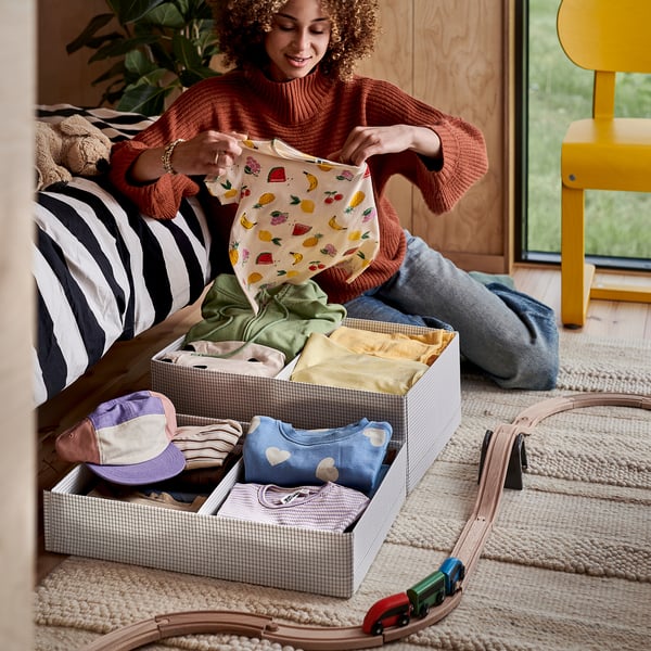 A person is organising baby clothes into a STUK storage box on a rug with a toy train.