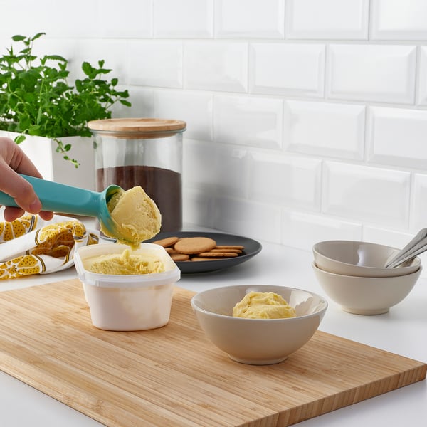Hand scooping yellow ice cream into bowl on wooden board with cookies and bowls, against white tiled wall.