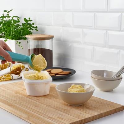 Hand scooping yellow ice cream into bowl on wooden board with cookies and bowls, against white tiled wall.