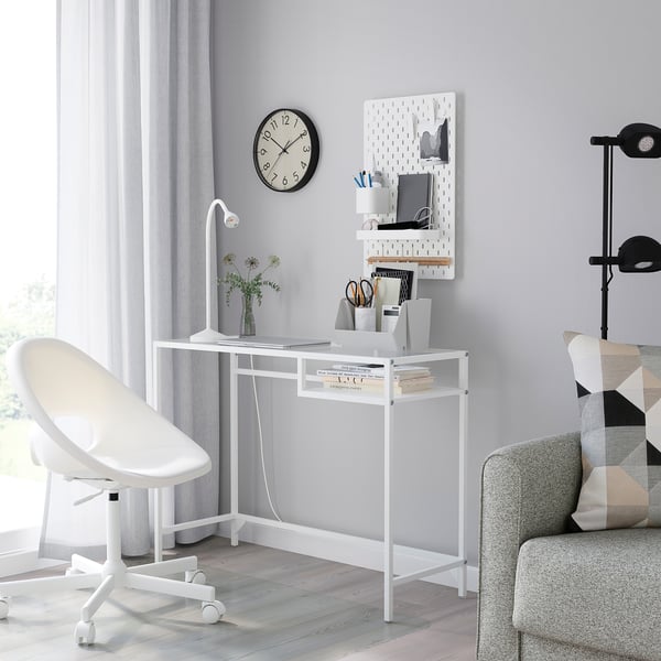 VITTSJÖ laptop table with glass top, metal frame, wall-mounted pegboard, white chair, grey sofa with pillows.