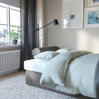 Minimalist bedroom with large bed, light blue blanket, and wall lamp. A plant by the window and white frames decorate the room.