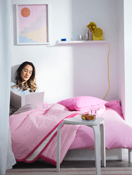 young woman sitting on bed with pink comforter in college dorm room reading a book
