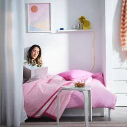 A young woman sitting on college dorm room bed with pink comforter reading a book
