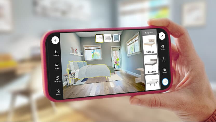 A hand holding a mobile device showing a scanned room with added IKEA dorm decor and furniture in IKEA Kreativ. The actual room is partially visible in the background.