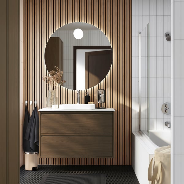 A modern bathroom with an ÄNGSJÖN wooden cabinet in dark brown. It a circular mirror and fixtures in silver. The sink area is sleek and minimalist.
