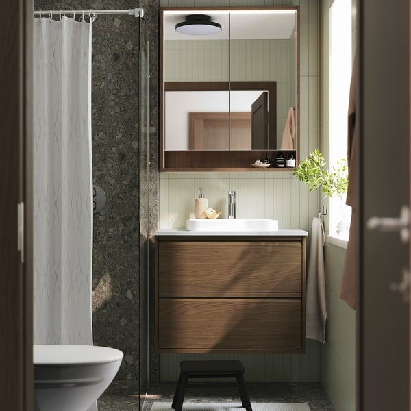 Modern bathroom with brown ÄNGSJÖN vanity, white sink, large mirror, green tiled walls, toilet, shower, black stool, towel