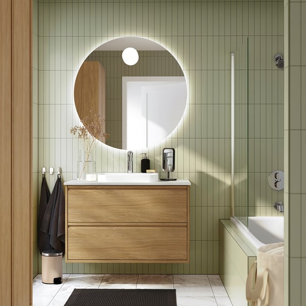 Modern bathroom with ÄNGSJÖN oak cabinet. Floating design, large round mirror, sleek sink, green walls, minimalist decor.