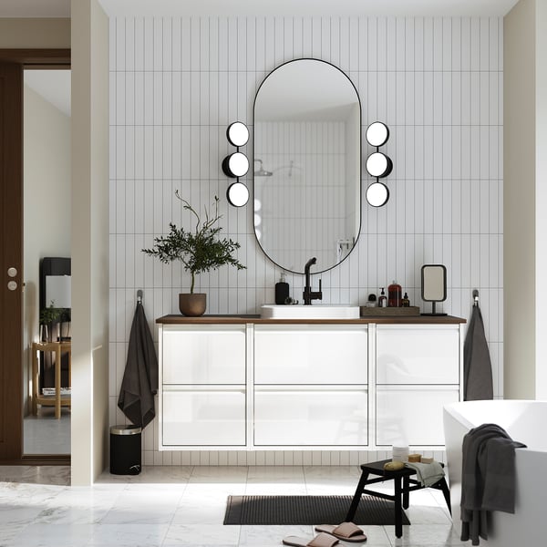Modern bathroom with ÄNGSJÖN vanity, glossy white drawers. Round mirror with lights, black accents. Plant, tub, door.