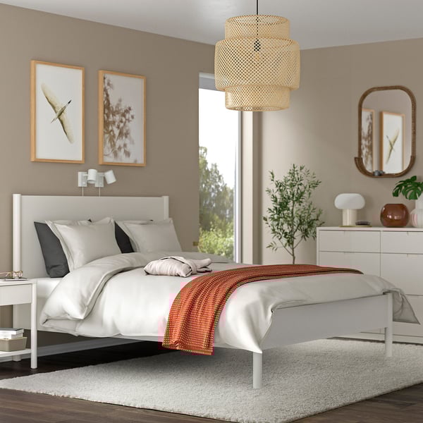 Bedroom with modern design; white bed, large rug under bed, hanging light, dresser with plants, framed art on walls.