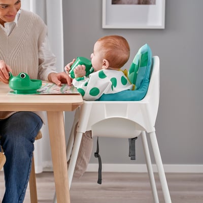 Baby in highchair reaches for toy, mum encourages.