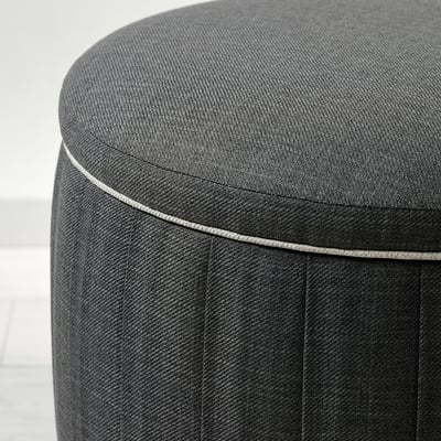 Small, round footstool with smooth, grey upholstery, subtle pleats, and detailed piping, showcasing quality craftsmanship.