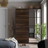 Bedroom with tall, mirrored AULI wardrobe, bed, chair, window, light fixture, linens, sandals, and plant.