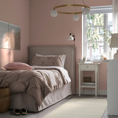 Cosy bedroom with BÄGARKLOCKA grey comforter on bed, pink walls, white bedside table, lamp, and window with curtains.